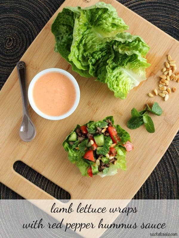 Overhead of items displayed on wooded cutting board: one lamb lettuce wrap, mint leaves, pine nuts, lettuce leaves, sauce and a spoon. Text overlay reads "lamb lettuce wraps with red pepper hummus sauce, rachelcooks.com"