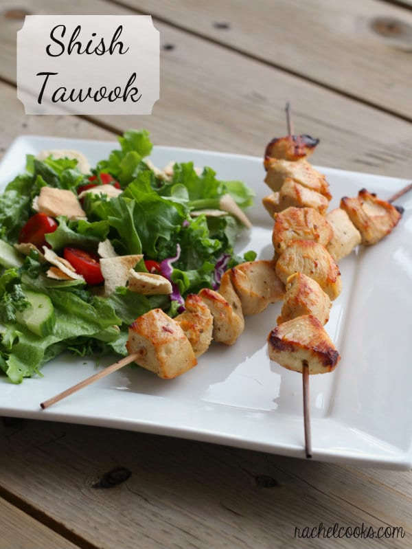 Two skewers with chicken and green salad on square white plate. Text overlay reads "Shish Tawook."