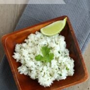 Overhead view of rice in a square wooden bowl, garnished with cilantro leaf and wedge of lime, on a gray cloth. Text overlay reads Cilantro-Lime Rice.