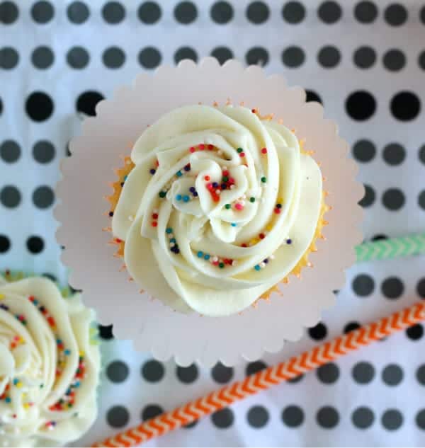 Overhead view of vanilla cupcakes on fancy scalloped paper round, with colorful straws, and polka dot background.