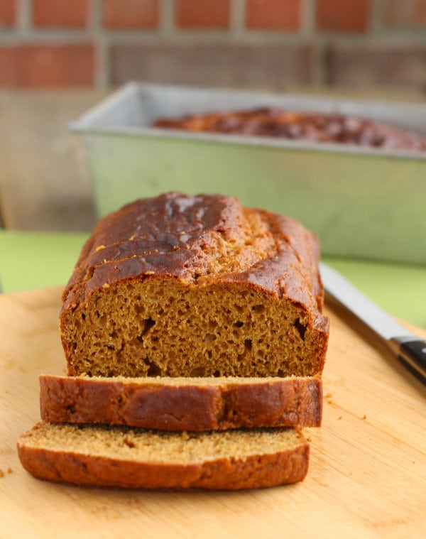 Front view of sliced loaf of pumpkin bread on wooden cutting board. In background is a loaf pan with pumpkin bread.