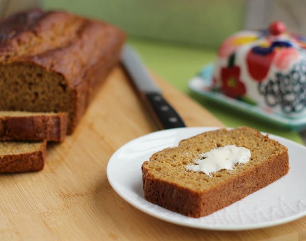 White plate containing slice of pumpkin bread with butter, sliced loaf in. background with bread knife, and fancy butter dish.