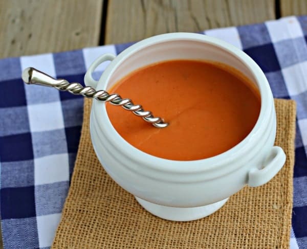 Image of white bean tomato soup in white bowl with fancy spoon, on burlap square. Blue checked fabric in background.