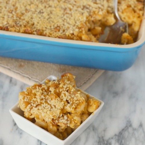 Pumpkin macaroni and cheese in a small white bowl, with a baking dish of macaroni and cheese in the background.