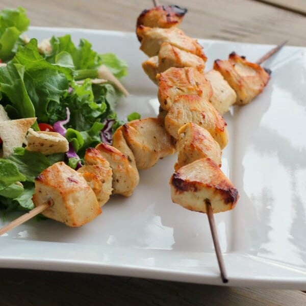 Two skewers with grilled chicken along with salad on square white plate.