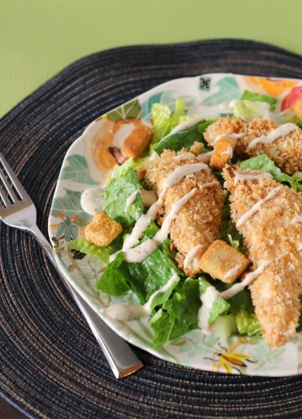 Partial image of chicken tenders served on a salad with dressing drizzled over all, on decorative plate with fork.