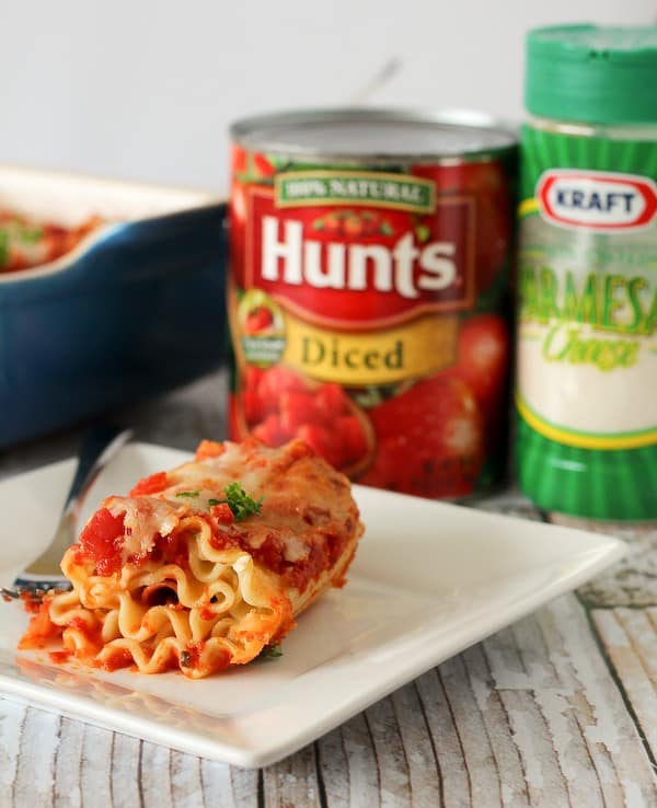 A rolled up lasagna noodle topped with tomato sauce, with a can of tomatoes and a package of parmesan cheese in the background.