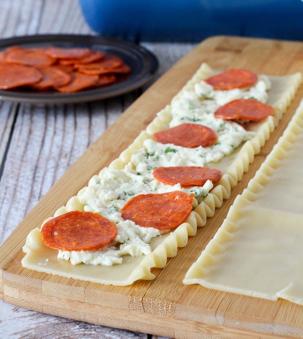 A cooked lasagna noodle laid out on a cutting board, topped with a ricotta cheese mixture and six pepperonis.