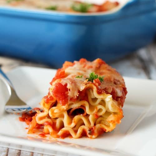 Lasagna pasta rolled up with pepperoni and cheese, coated in a tomato sauce. It is on a white plate and a blue baking dish appears in the background.