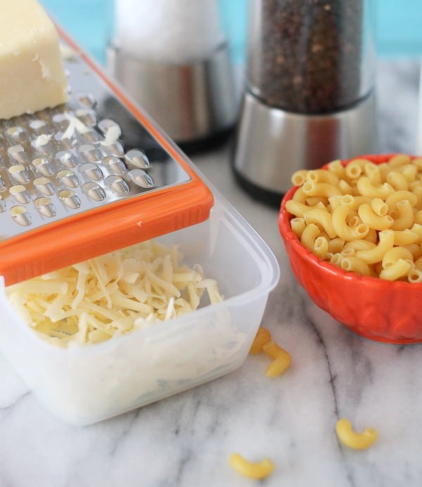 White grated cheese in a box grater. An orange bowl of uncooked macaroni sits nearby, salt and pepper shakers are in the background. 