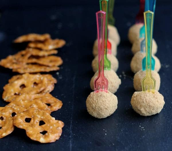 Two rows of cinnamon cheesecake balls with brightly colored forks inserted upright, and a row of pretzel flats, on black background.