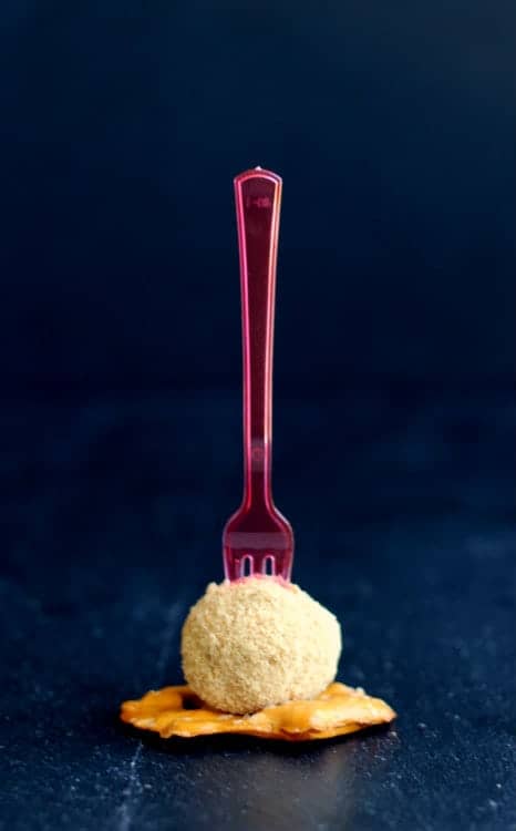 A cinnamon cheeseball on a flat pretzel, with pink plastic fork inserted, on black background.