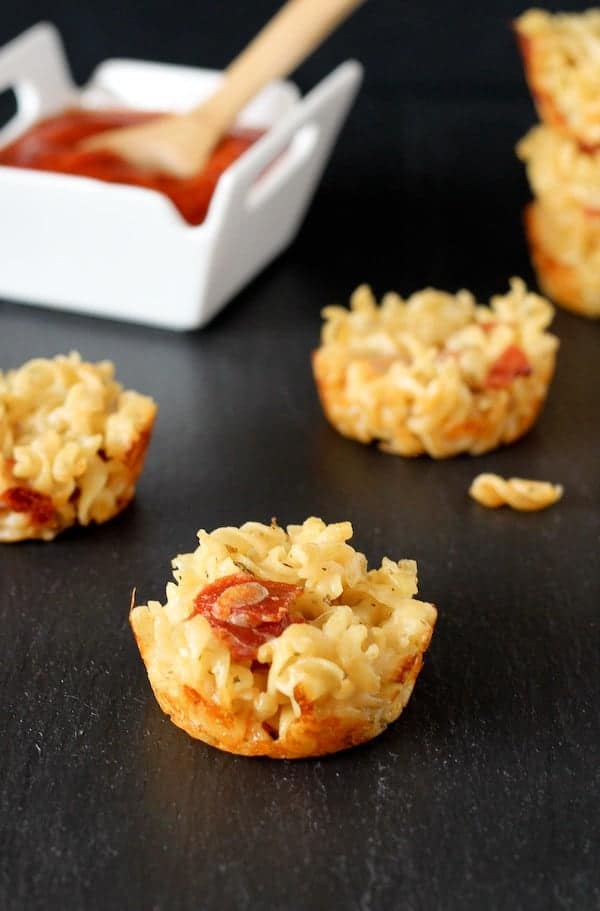 Front view of several mac and cheese muffins, with white bowl containing pizza sauce and wooden spoon in background.