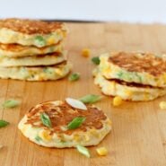 Several corn cakes on wooden cutting board, garnished with sliced green onions and corn kernels.