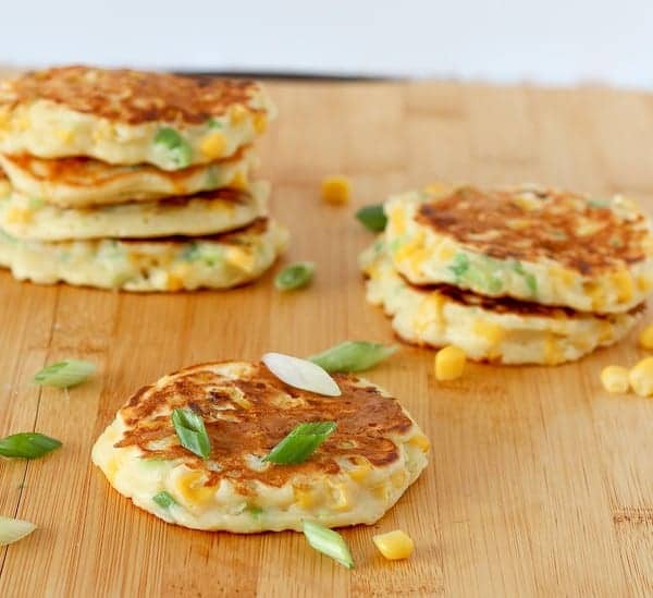 Several corn cakes on wooden cutting board, garnished with sliced green onions and corn kernels.