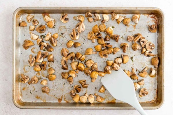 A white spatula stirs roasted white mushrooms on a baking sheet.