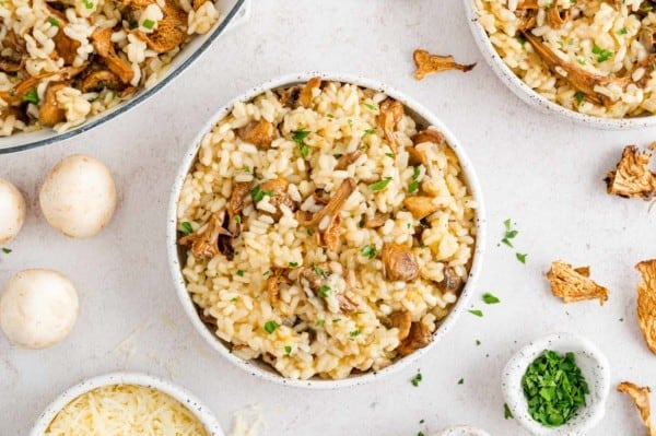 Overhead view of a bowl of mushroom risotto surrounded by scattered mushrooms and small bowls of parsley and parmesan.