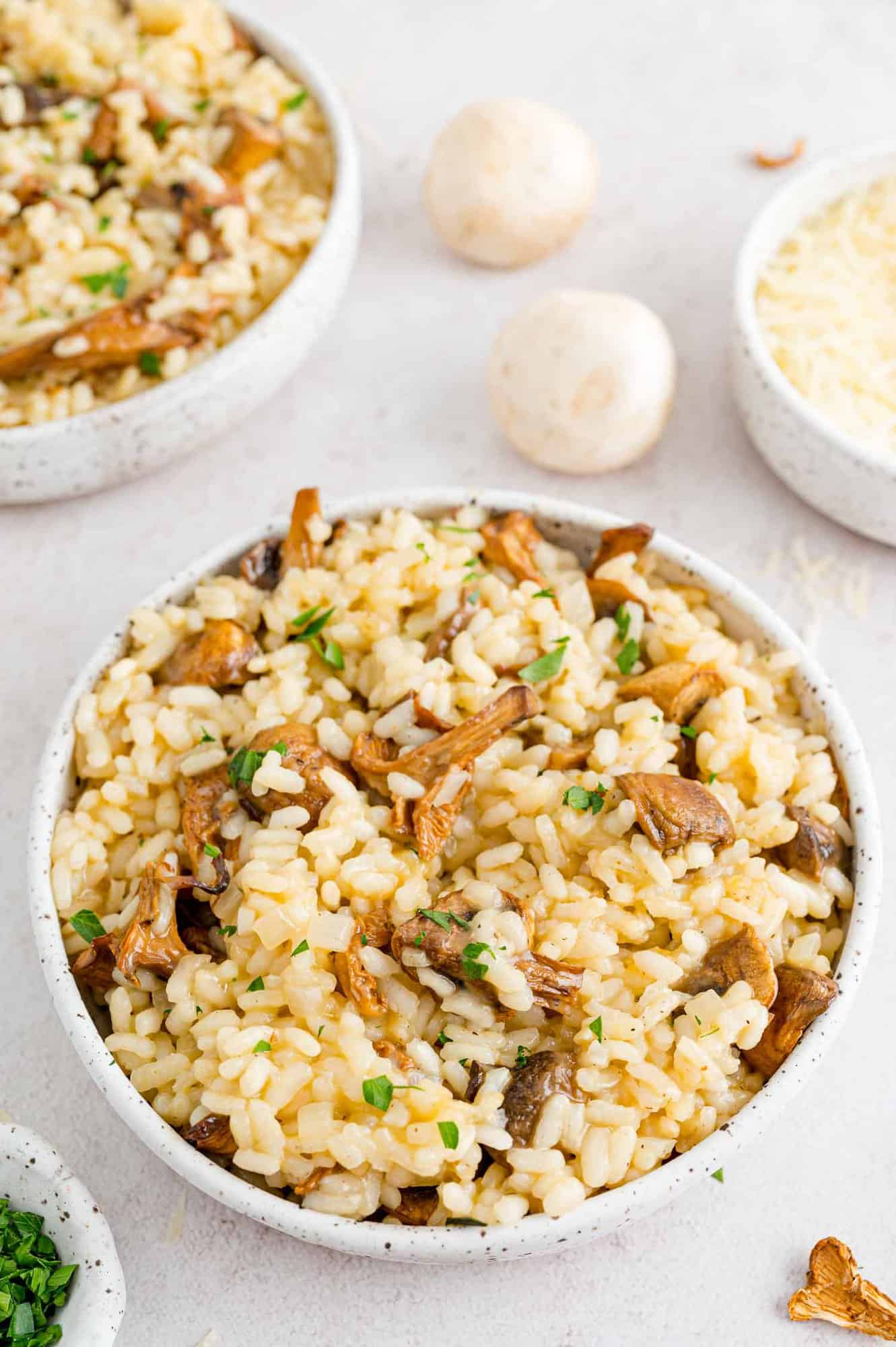 A bowl of mushroom risotto next to scattered whole white mushrooms and a second bowl in the background.