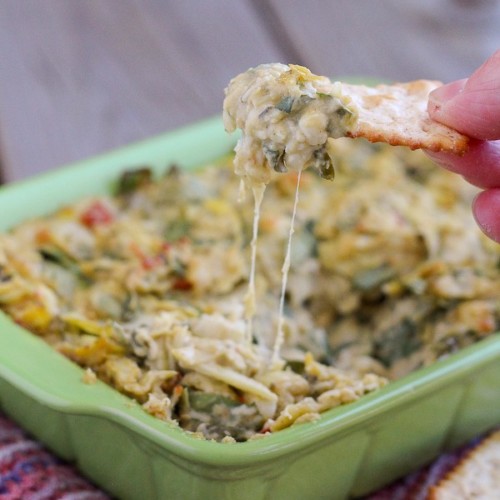 Front view of casserole dish containing artichoke dip with finger holding cracker loaded with dip.