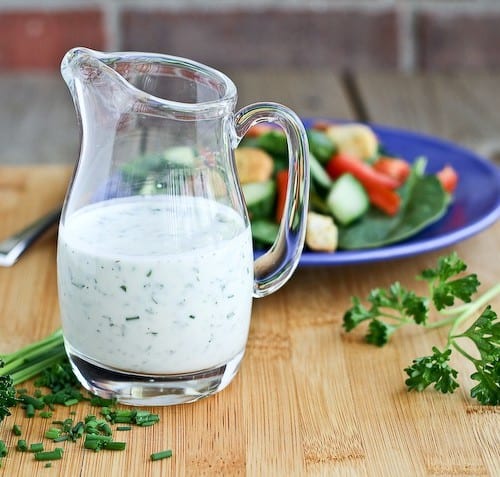 Small clear glass pitcher of ranch dressing with salad in background.