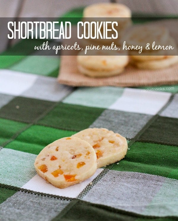 Two shortbread cookies on green plaid cloth, with additional cookies in background. Text overlay reads "Shortbread cookies with apricots, pine nuts, honey & lemon."
