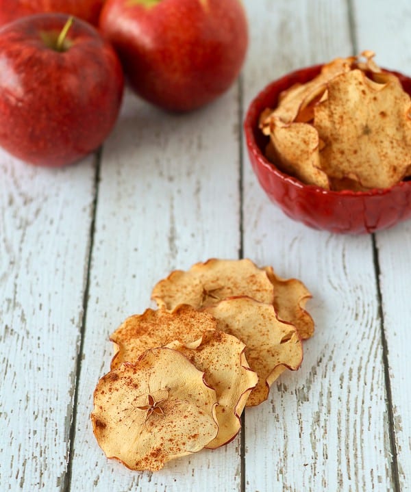 Several apple chips stacked on white board background, with red bowl containing more apple chips and a few apples in the background.