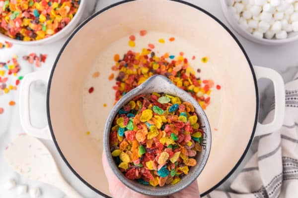 A cup of Fruity Pebbles cereal held over melted marshmallows in a saucepan.