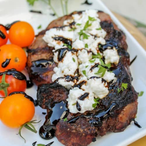 Goat cheese topped steak with balsamic drizzle, with tomatoes and parsley on white plate.