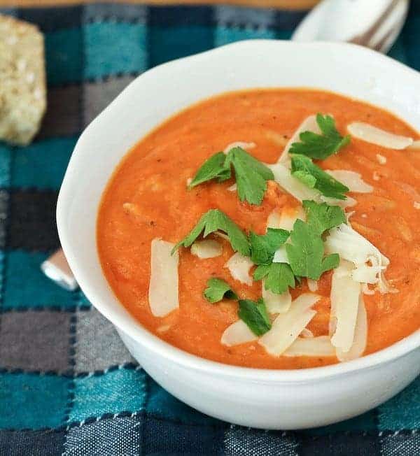 Image of creamy tomato soup in a white bowl, garnished with parsley leaves and shredded parmesan cheese. Soup is pictured on plaid blue cloth with spoon nearby.
