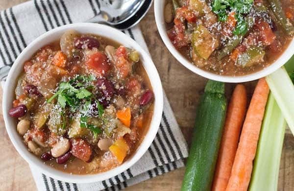 Image of Slow Cooker Minestrone with Quinoa in 2 white bowls on black and white striped fabric. Also included are a zucchini, 2 carrots, and 2 stalks of celery.