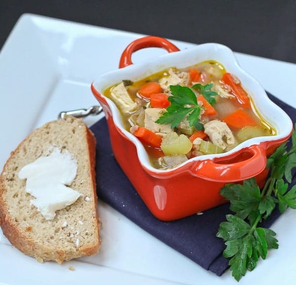 Soup in decorative orange bowl, on square white plate with buttered bread, spoon, and sprig of parsley.