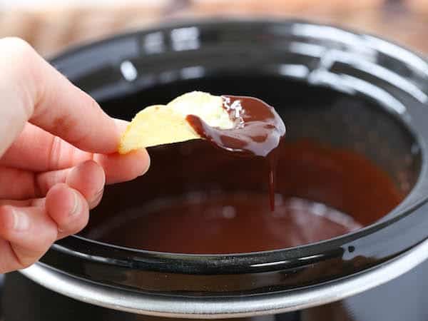 Hand holding potato chip dipped in chocolate fondue with slow cooker in background.
