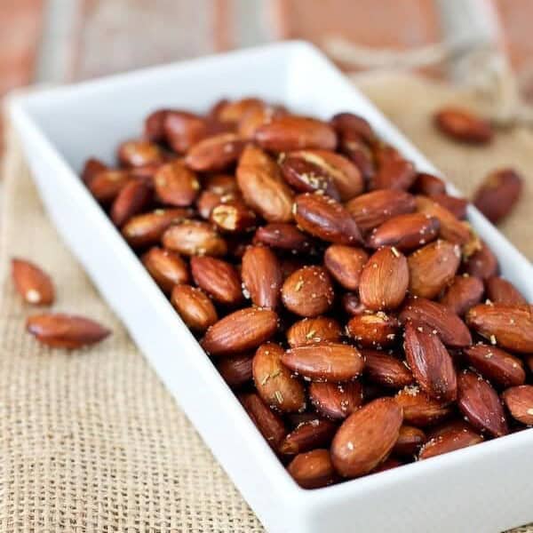 A rectangular white dish filled with rosemary roasted almonds sits on a piece of burlap, with a few almonds scattered around.