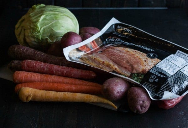 Image contains package of corned beef, several rainbow colored carrots, red skin potatoes, and a head of green cabbage.