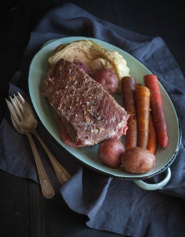Overhead view of oval platter of corned beef brisket dinner, on a blue cloth with antique silver fork and spoon along side.