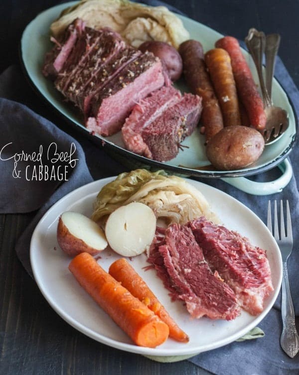 A white plate, containing corned beef slices, 2 carrots, red skinned potato cut in half, and cabbage. Included in the image is oval platter with remaining sliced corned beef, vegetables, with serving utensils. Also in the image is a text overlay that reads " corned beef & cabbage."