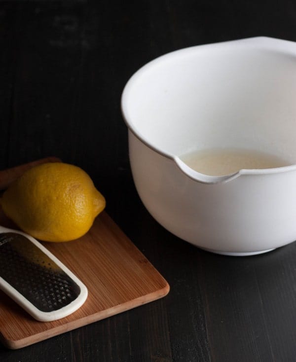 Image consists of partially shown white mixing bowl, cutting board, whole lemon, and zester, on a black background.