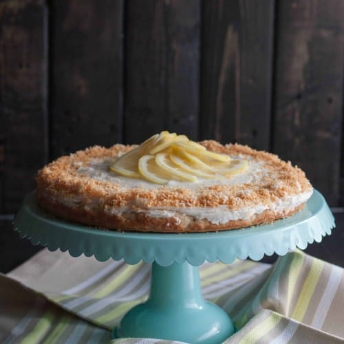 Coconut lemon cake displayed on pedestal cake stand, with striped cloth underneath and dark background.