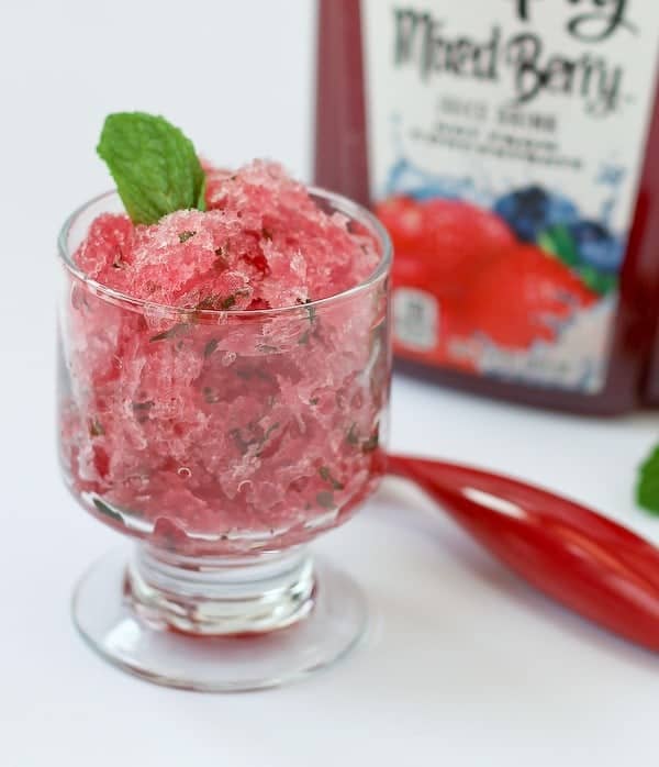 Image of granita with rum in a clear glass dessert dish, garnished with mint leave, with red spoon and Simply Mixed Berry in background.