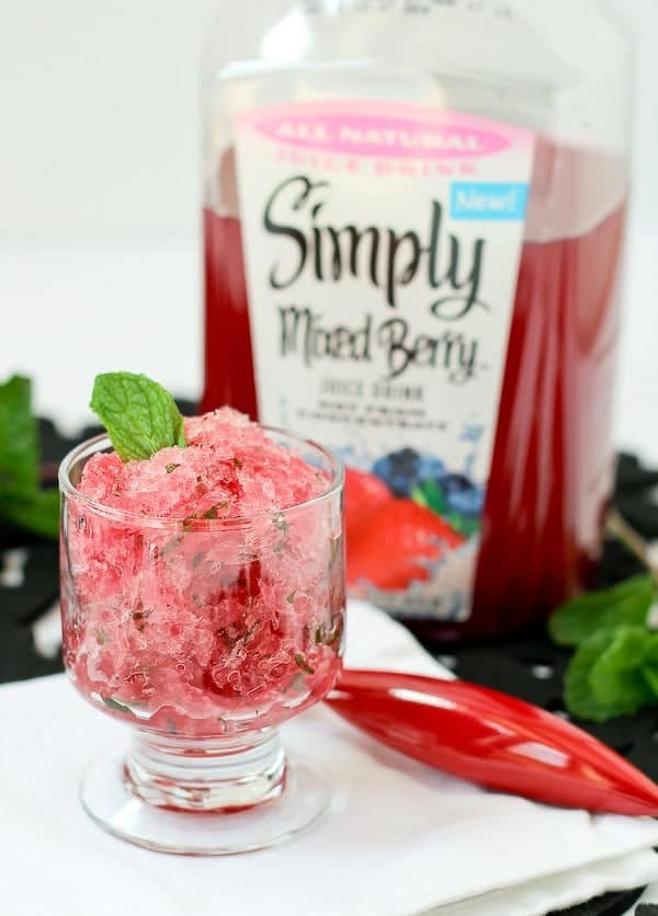 Image consists of footed clear glass dessert dish with icy red granita, garnished with single mint leaf. A plastic bottle of Simply Mixed Berry juice is in the background.