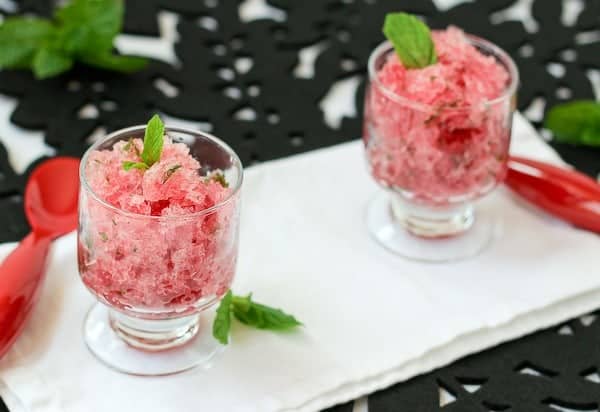 Two clear glass dessert dishes of granita with rum on a white serving tray, with 2 red spoons. Mint leaves are scattered around the desserts.