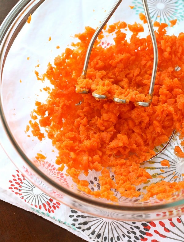 Clear glass mixing bowl of mashed cooked carrots with metal potato masher.