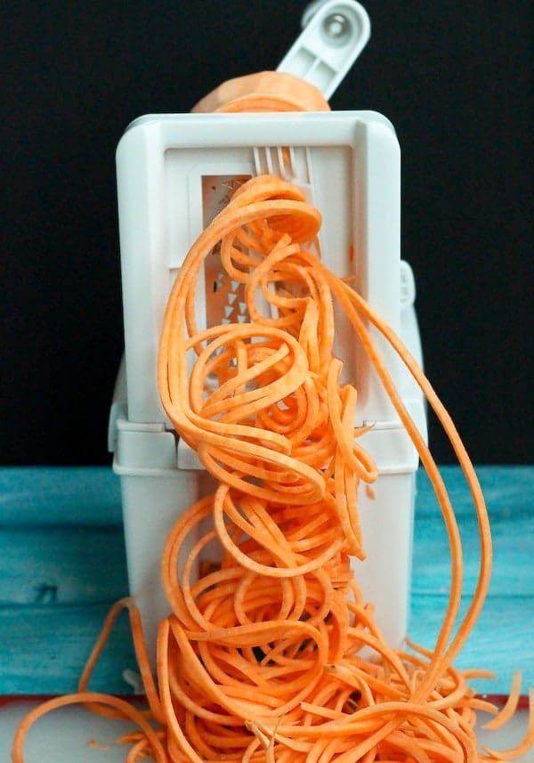 Image of sweet potato noodles emerging from the blades of a spiralizer, a bright turquoise background.