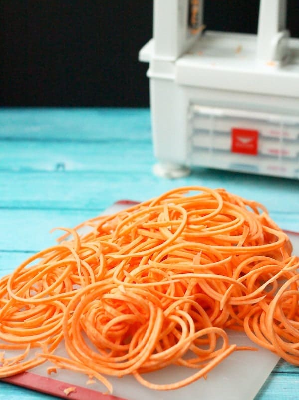 In the foreground, a pile of sweet potato noodles rests on an acrylic cutting board, with a partial picture of a spiralizer in the background.