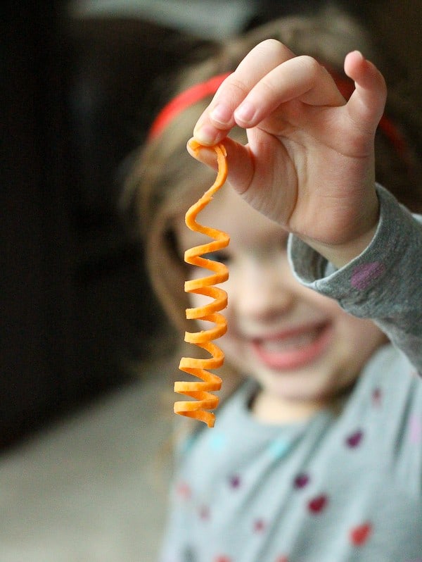The image of a young child holding a single sweet potato noodle between her thumb and first finger. 