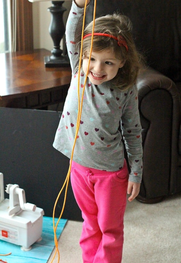 Pictured is a toddler girl, in a living room, holding a very long straight sweet potato noodle, demonstrating how long it is--taller than she is!