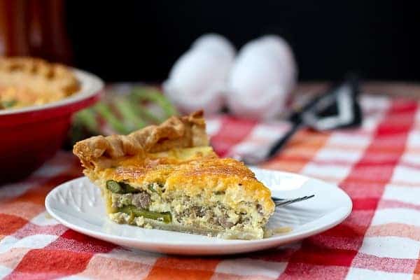 Slice of quiche on a round white plate. Eggs and fresh asparagus in the background.
