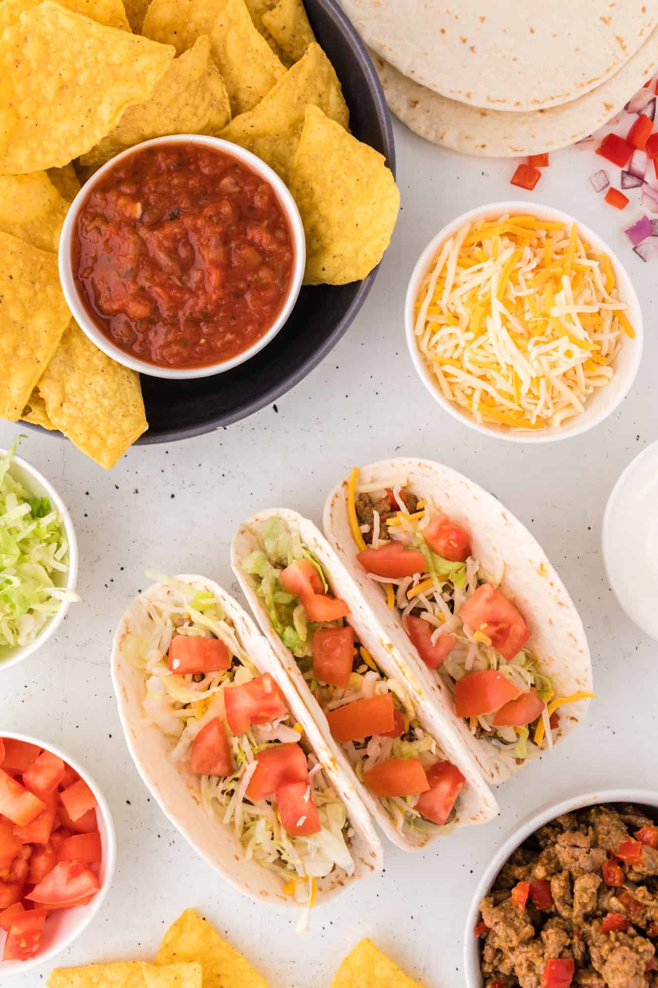 Overhead view of a taco night spread including three assembled turkey tacos, a plate of tortilla chips and salsa, and bowls of various taco toppings.