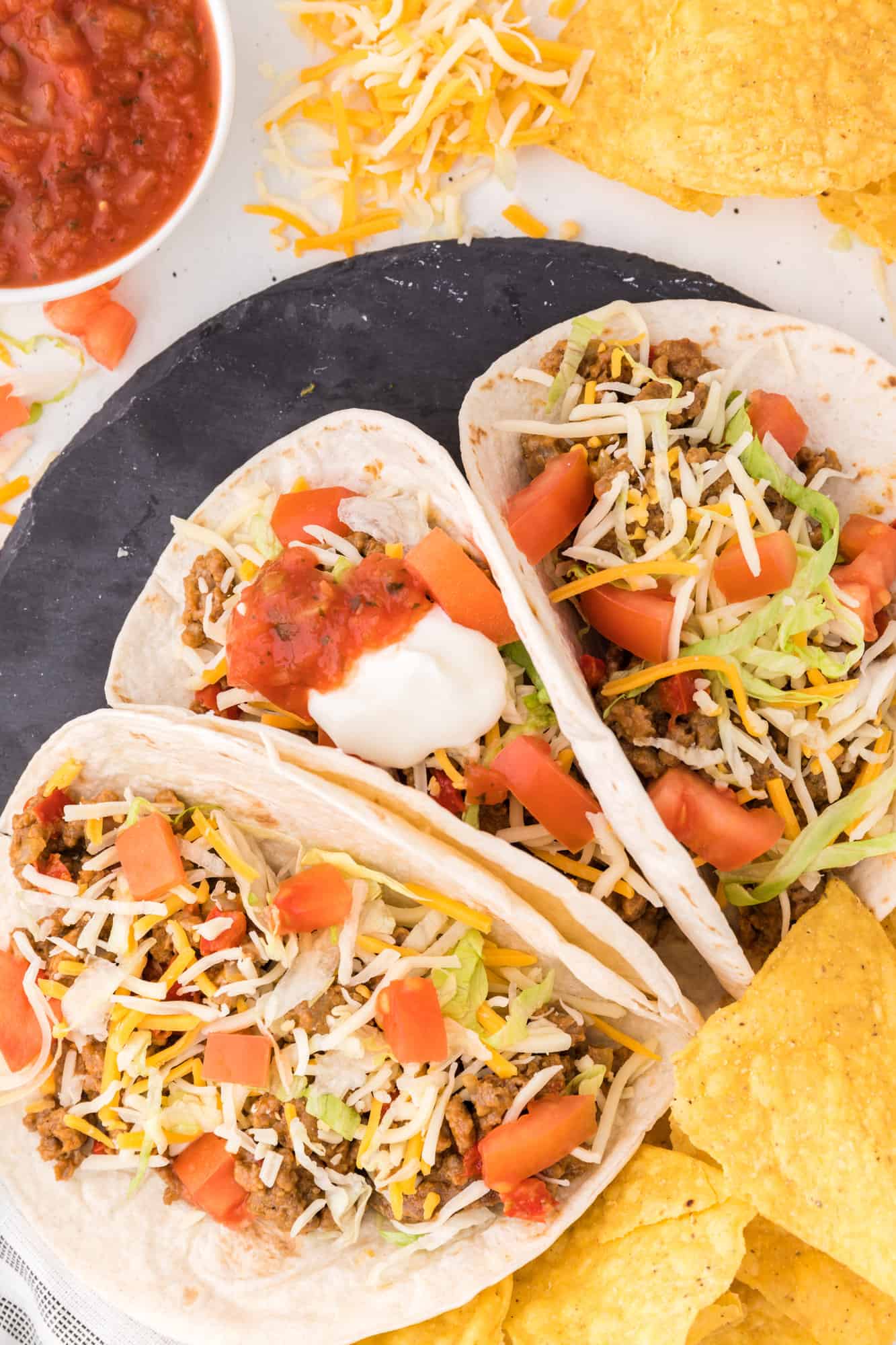 Overhead view of three turkey tacos on a black stone plate next to tortilla chips and salsa.