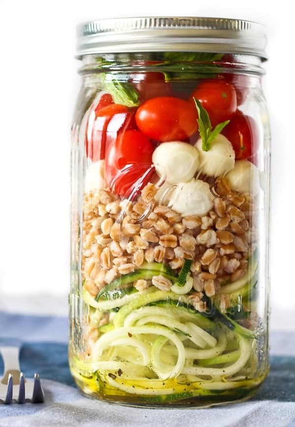 Image of Mason jar with layers, beginning at the bottom, of vinaigrette, zucchini noodles, farro, mozzarella pearls, grape tomatoes and basil leaves. Also pictured is a fork.